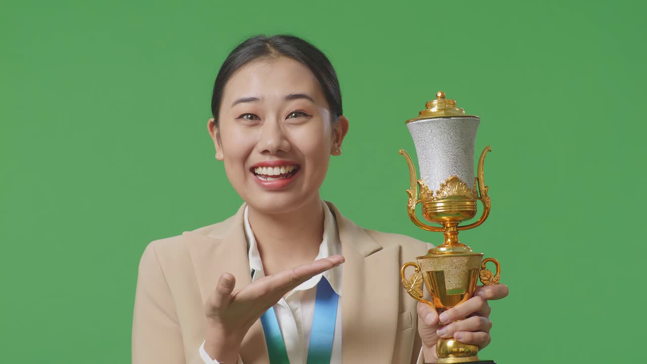 손에 금메달을  슈트를 입은 아시아 사업가 여성은 손에 금 트로피를 가리키며 미소 짓고 스튜디오의 녹색 스크린 배경에서 첫 번째 우승자로 자랑스럽게 승리합니다.