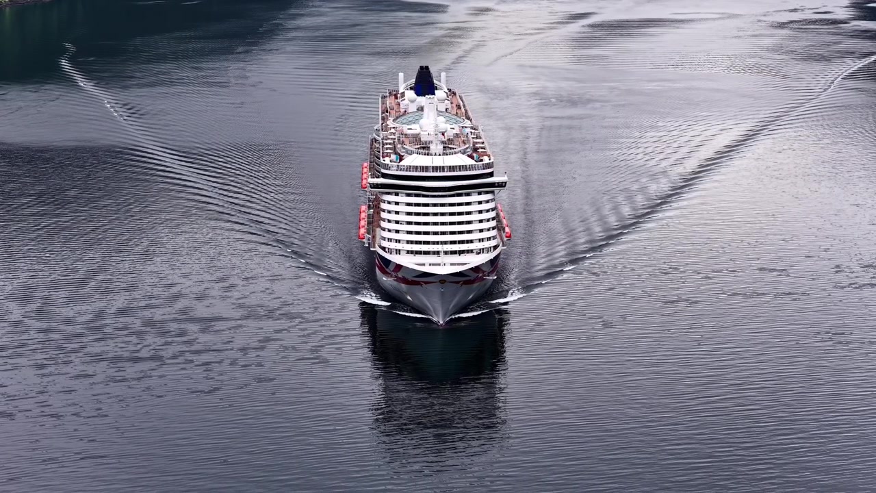 Stort krydstogtskib sejler fremad på rolige fjordvande og skaber krusninger i Norge.
