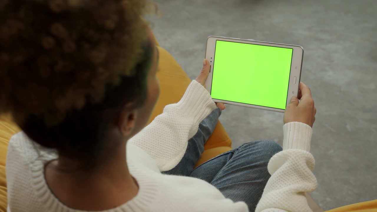 A black curly-haired woman uses a tablet device with a chromakey while sitting on the couch
