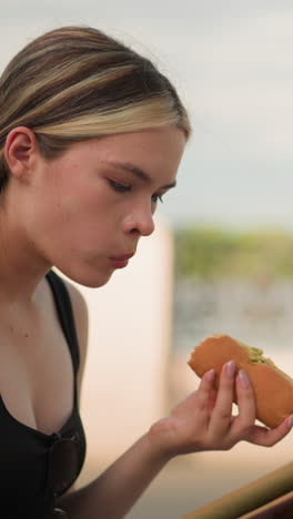 mujer con camiseta negra sentada al aire libre, disfrutando de una hamburguesa mientras lee un libro, con una valla decorativa y un cielo claro y brillante en el fondo, participando en la lectura casual y comiendo al aire libre