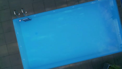 vista aérea ascendente y giratoria de una piscina con agua azul brillante rodeada por una cubierta de piedra gris, paisaje verde de una casa moderna y lujosa
