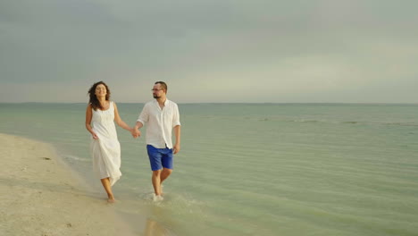 Joven-Pareja-De-Enamorados-En-Ropa-Ligera-Sin-Preocupaciones-Corriendo-En-Una-Playa-Tropical-Limpia