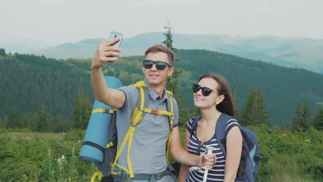 A-Young-Couple-Of-Tourists-With-Backpacks-Take-Pictures-Of-Themselves-With-A-Smartphone-On-A-Beautif