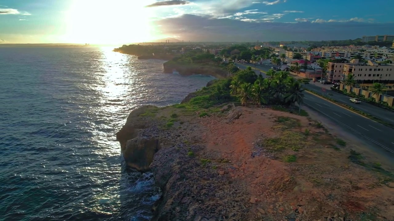 Ripresa aerea diurna con drone che si sposta a sinistra sopra il traffico dell'autostrada costiera e le scogliere rocciose per rivelare la costa del Mar dei Caraibi durante il tramonto a Santo Domingo, Repubblica Dominicana