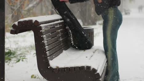 partial view of lady removing guitar pack to place on snow-covered bench in snowy park, with blurred figures walking in distant background surrounded by frosted trees and serene winter atmosphere