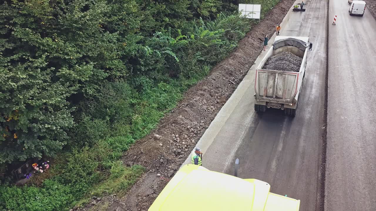 A truck with bitumen slowly drives up to a paver on a highway near a forest landing on the background of road repair works near the city. Aerial view.