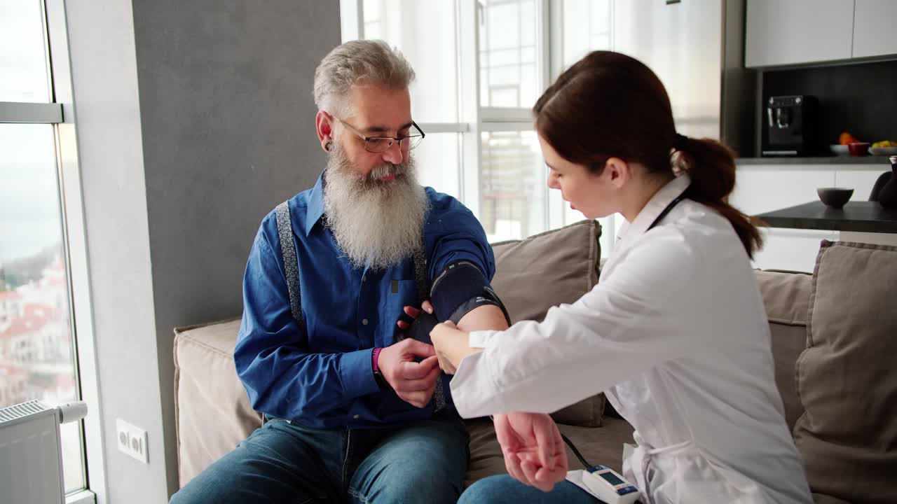 A brunette doctor girl in a white coat examines an elderly man with gray hair and a lush beard in a blue shirt using a heart rate monitor and measures the mans blood pressure in a modern apartment