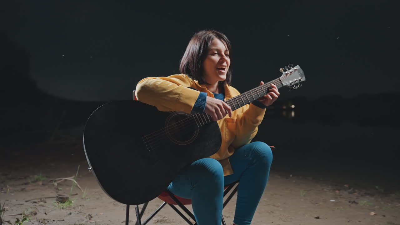 Festa all'aperto energica con donna che canta. Vivace scena di falò con chitarrista asiatica sorridente che si esibisce. Musicista allegra che intrattiene gli amici attorno al fuoco riscaldante sotto il cielo notturno.