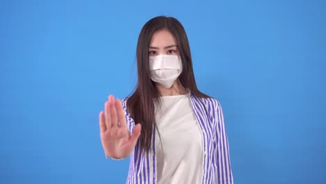 beautiful young asian woman with a protective medical mask on her face shows a stop gesture while standing on a blue background