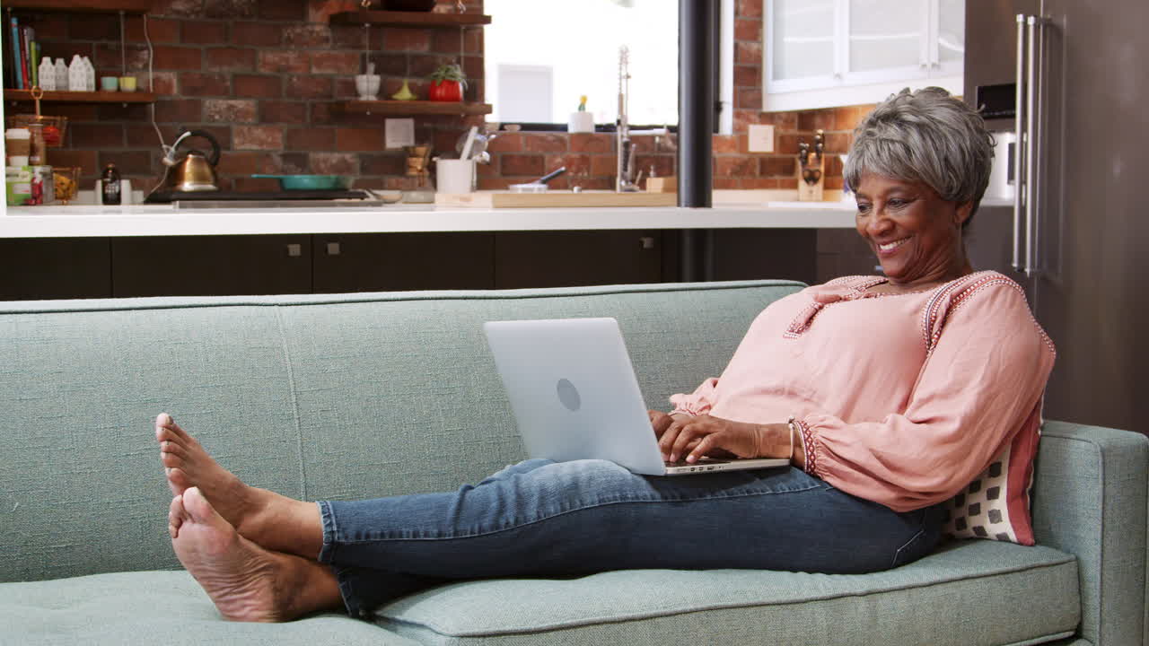 Senior Woman Relaxing On Sofa At Home Using Laptop To Shop Online