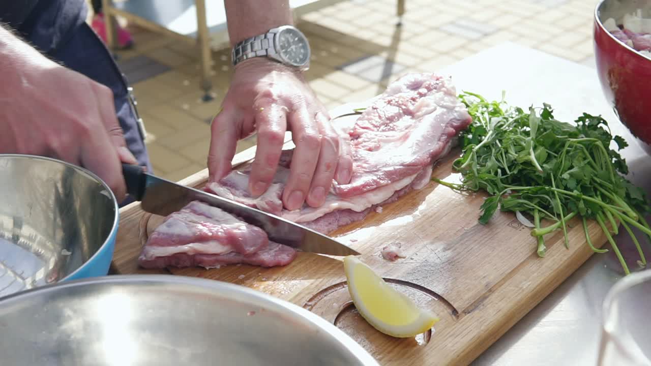 Person's hands cutting a piece of raw meat using a butcher's knife on a wooden surface outside. Barbeque preparation. shot in 4k