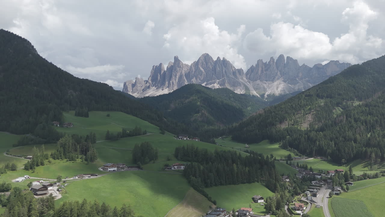 Aerial drone view of the Dolomite Alps with lush green valley, forested hills and dramatic mountain peaks under a cloudy summer sky