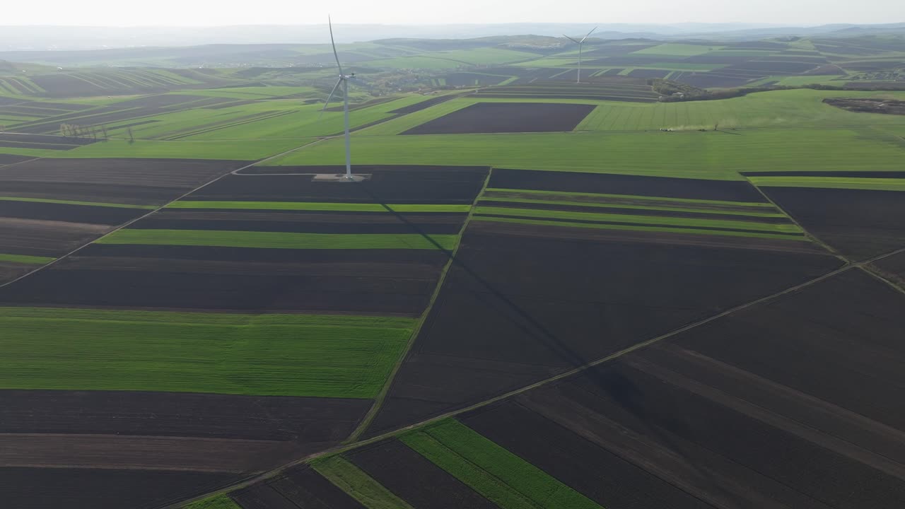 Green and brown fields with a wind turbine casting a long shadow, aerial view