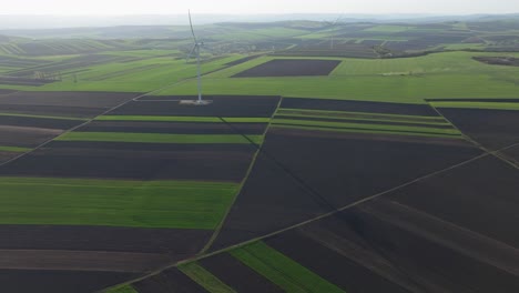 Green-and-brown-fields-with-a-wind-turbine-casting-a-long-shadow,-aerial-view