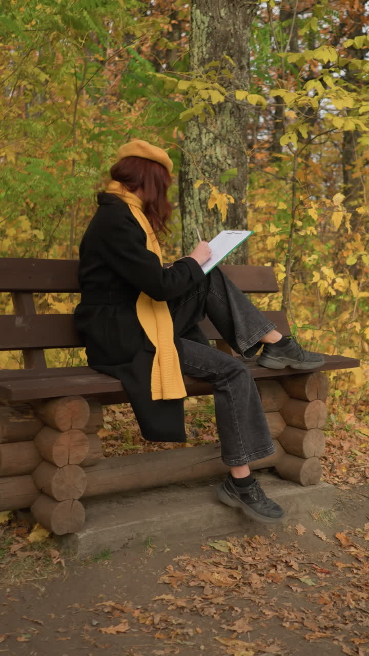 vista trasera de una dama dibujando sentada al aire libre en un banco de madera en soledad, rodeada de follaje de otoño, inmersa en la creatividad, con abrigo negro y bufanda amarilla mostaza