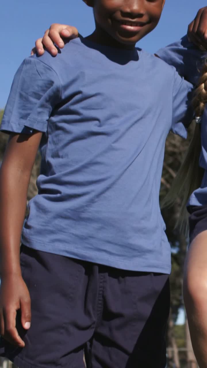 Vídeo vertical: Menino se ajeitando em campo de parque, menina entrando, dando o braço e posando com camisetas azuis claras