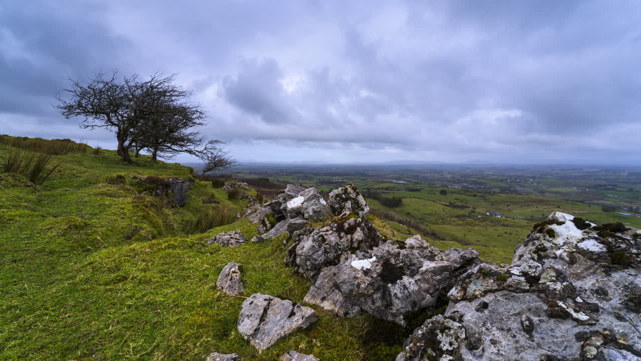 Panorama-timelapsevideo af et landskab med et enkelt træ og stengærde i en græsmark, bakker og en sø i horisonten på en skyet dag, set fra Carrowkeel i County Sligo i Irland