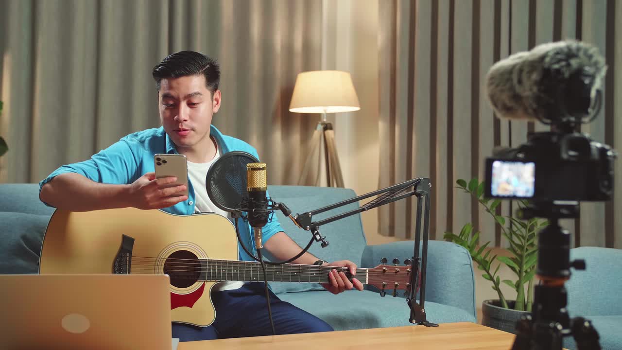 Asian Man Is A Vlogger With Guitar Read Comment On Mobile Phone. The Male Is Streaming For His Social Media Channel. The Man Is Broadcasting Live On The Internet.