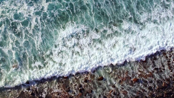 Ripresa dall'alto con drone di onde oceaniche turchesi che si infrangono su una spiaggia coperta di piccoli ciottoli, con una splendida schiuma bianca