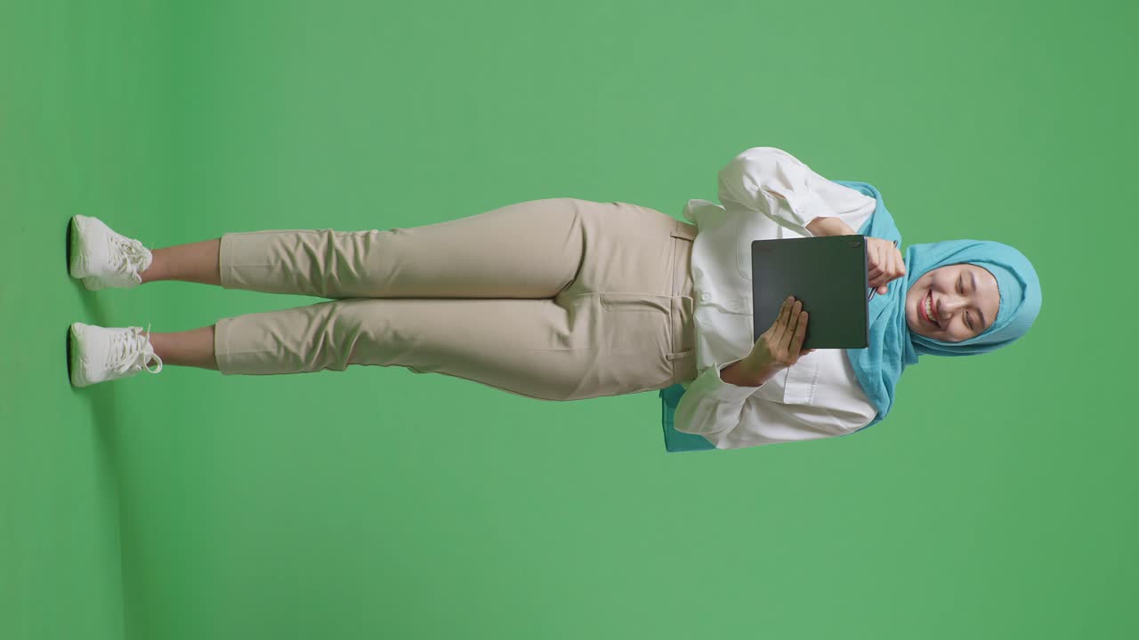 cuerpo lleno de mujer musulmana asiática sonriendo y tomando nota en una tableta mientras estaba de pie en el estudio de fondo de la pantalla verde