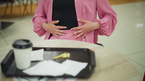 lady in pink dress gently rubbing her stomach with two hands after a meal, while her left hand rests on the table, a tray with a coffee cup, napkins, and leftover fries lies in front of her