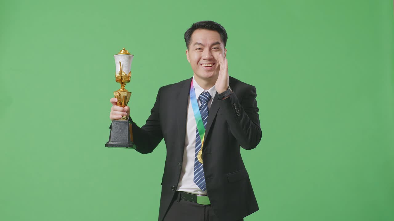 슈트와 타이를 입은 아시아 사업가, 금메달을 들고, 금 트로피를 들고, 스튜디오에서 녹색 화면 배경에 입을 고 긍정적으로 소리쳤다.