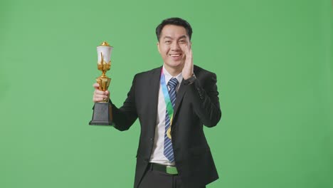 슈트와 타이를 입은 아시아 사업가, 금메달을 들고, 금 트로피를 들고, 스튜디오에서 녹색 화면 배경에 입을 고 긍정적으로 소리쳤다.