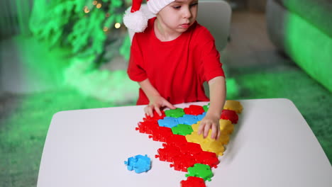 child makes festive puzzle. a child in a red shirt and santa hat assembles a puzzle on a white table amid festive decorations