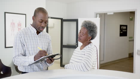 African-american-male-doctor-taking-notes-and-talking-with-senior-woman-in-hospital,-slow-motion