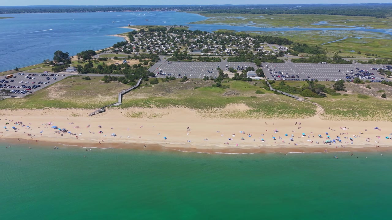 Panning drone fly of crowded Salisbury Beach lines a calm green Atlantic Ocean with rows of umbrellas, dunes, and large parking lots behind, Massachusetts, United States
