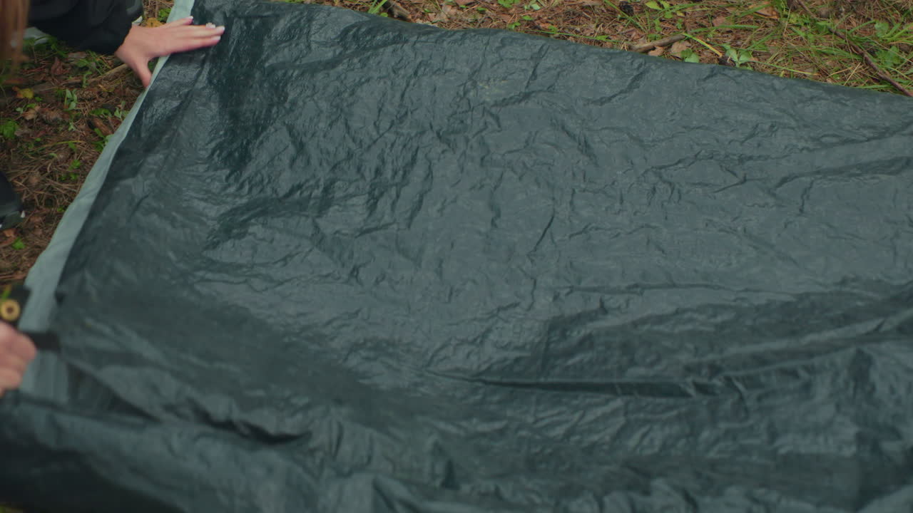Close up of lady unpacking red and gray tent on ground in forest while friend assists beside her, both leaning in close during outdoor camping activity surrounded by dry leaves and green undergrowth