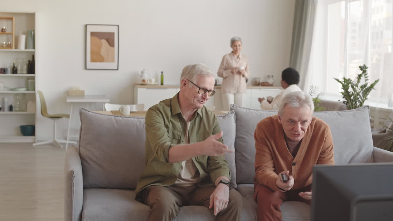 Medium long of two senior Caucasian men sitting on couch in living room, having discussion in front of TV-set