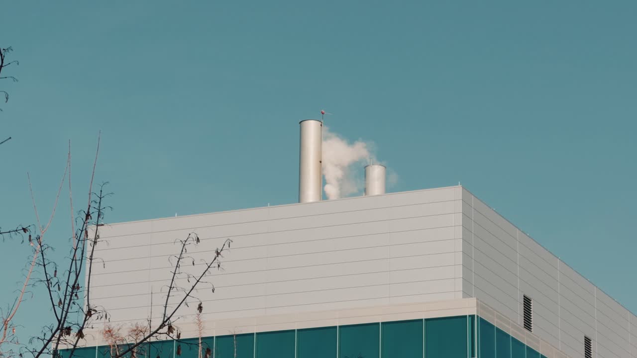 Inquadratura cinematografica stabile di vapore che si diffonde dai comignoli sui tetti di un edificio industriale moderno