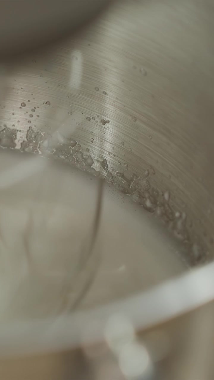 Close up of spoon stirring white liquid in metal bowl. Interior shows water droplets on stainless steel surface. Spoon movement is steady with slight angle changes emphasizing