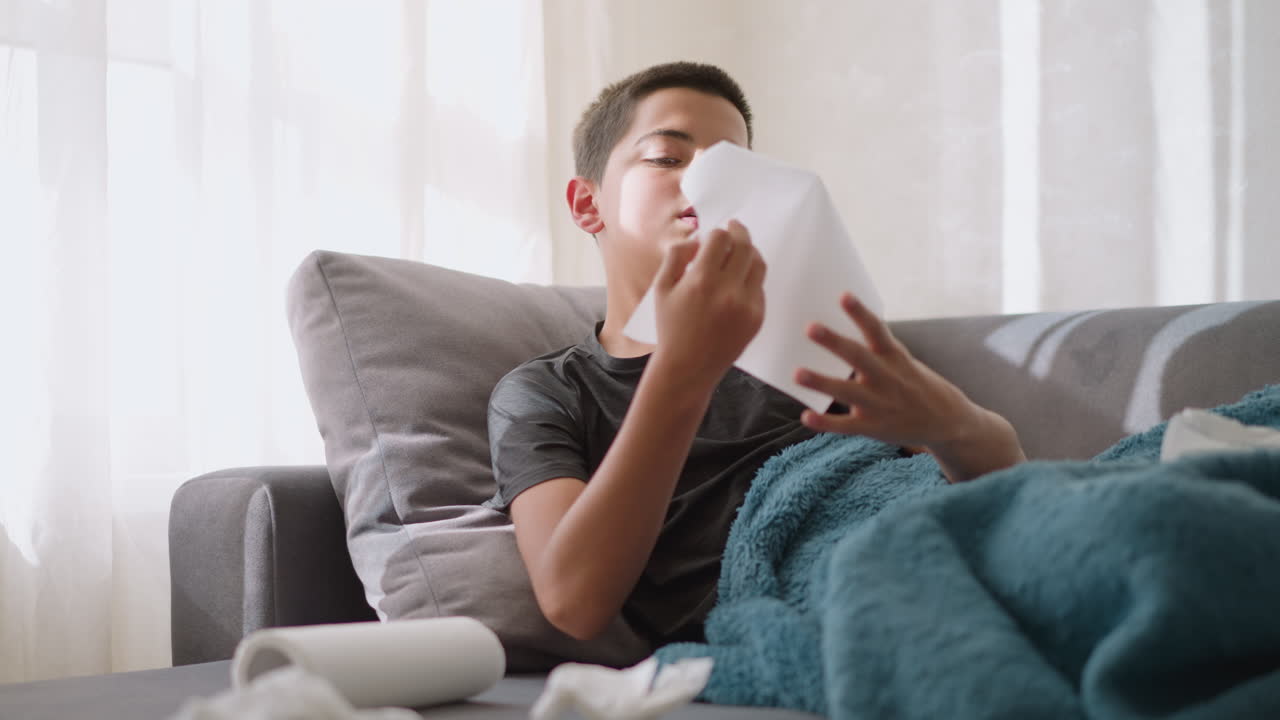 Sick boy lying on couch gets napkin to sneeze into it while surrounded by used napkins on the table and couch, as he rests and recovers from flu or cold at home