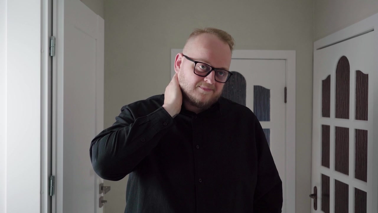 Middle-aged man with glasses rubs the side and back of his neck indoors, wincing as if from muscle tension or strain.