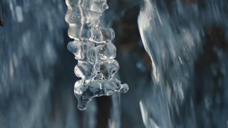 drop falling on icicle in a frozen river with flowing water in background, close up, slow motion