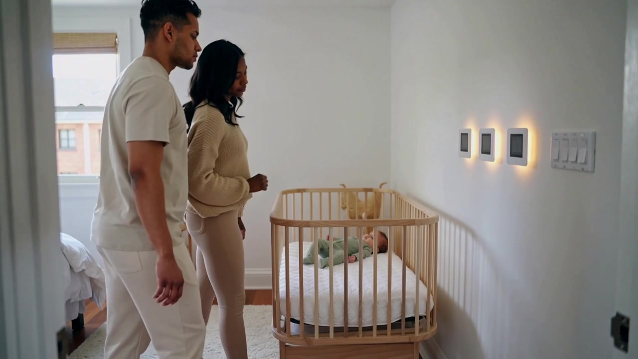 Entering doorway, couple in beige clothes touching lit panels in nursery, checking baby in crib