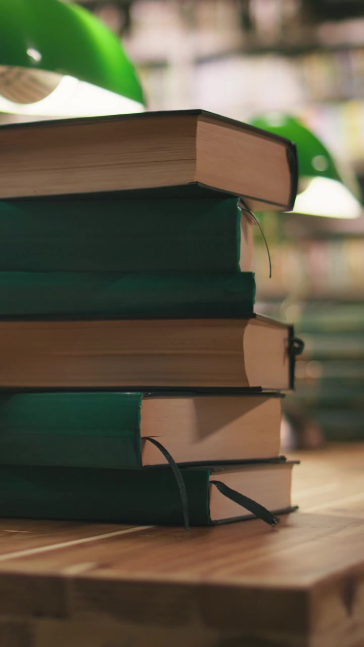 Video verticale: Primo piano di libri verdi impilati su una scrivania in biblioteca sotto lampade verdi.