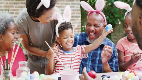 happy african american son painting easter eggs with grandparents and family in garden, slow motion