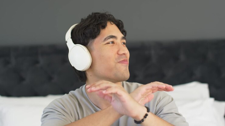 Uomo asiatico a letto che ascolta musica con cuffie bianche, alzando le dita, formando una X, mimando il testo.