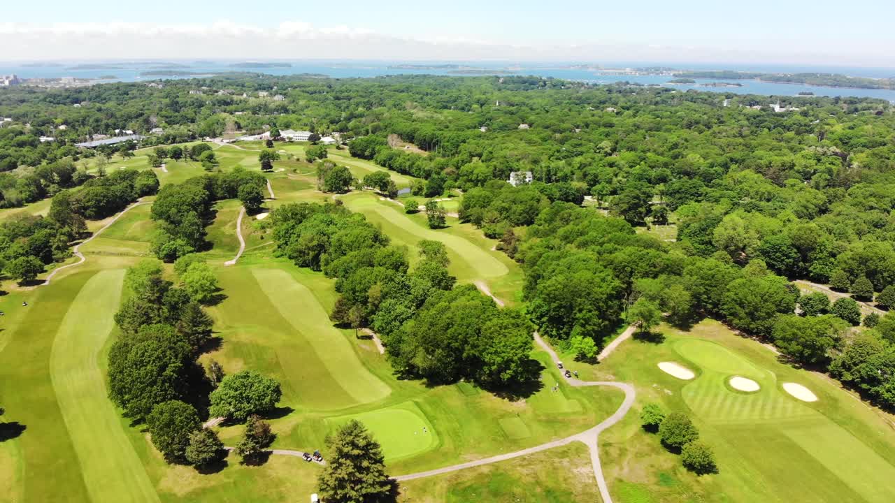 드론이 섬을 보여주는 항구나 만을 향해 날아가는 골프 코스의 주간 조감도, 골퍼들은 아래의 푸른 잔디에 점을 찍고 나무는 풍경에 점을 찍습니다.