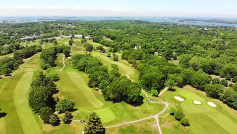 a daytime aerial view of a golf course as the drone flies toward the harbor or bay showing islands, golfers dot the green grass below and trees dot the landscape