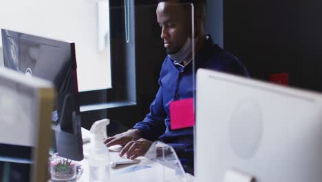 hombre afroamericano con máscara facial bajada usando computadora en una oficina moderna