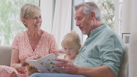 Abuelos-Sentados-En-El-Sofá-Con-Su-Nieta-En-Casa-Leyendo-Un-Libro-Juntos