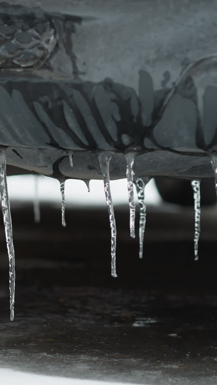 Close-up of car's fog light encased in ice with hanging icicles, displaying harsh winter effects on vehicle exteriors and a blurred red car in the background