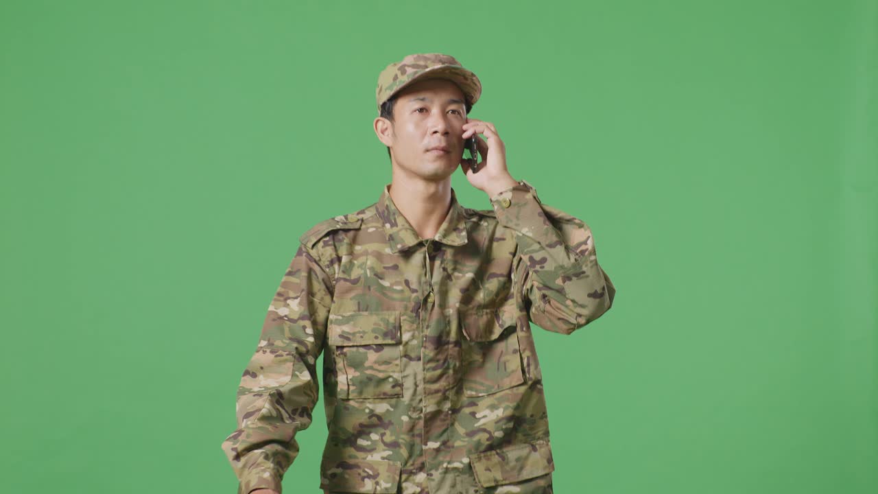 아시아 남자 군인이 스마트폰으로 이야기하고 녹색 스크린 배경 스튜디오에서 는 동안 주위를 둘러보고 있습니다.