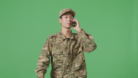 soldado hombre asiático hablando en el teléfono inteligente y mirando a su alrededor mientras camina en el estudio de fondo de la pantalla verde