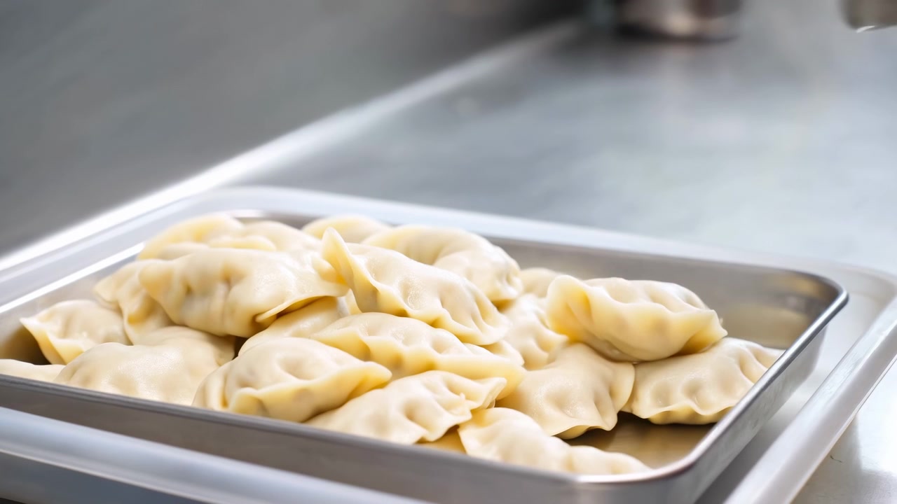 Plump uncooked dumplings with pale glossy dough and neat pleated crimps gather inside rectangular stainless steel tray on kitchen counter while more pieces descend from above, creating fresh