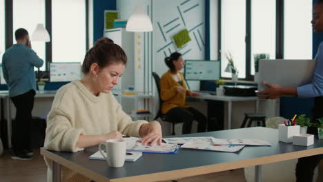 African-american-woman-holding-laptop-asking-colleague-looking-at-clipboard-with-charts-for-advice-regarding-financial-data